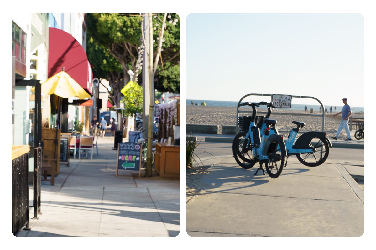 A Main Street sidewalk narrowed by cafe railings, planters, and a sandwich-board sign, and two rental bikes parked across the beach path beside a crosswalk sign.