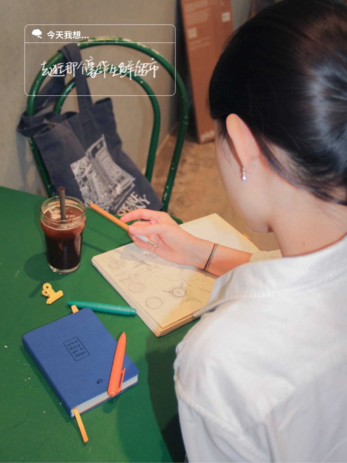A social post: someone drawing at a green cafe table with an iced coffee and the journal beside them, overlaid with the day's intention module.