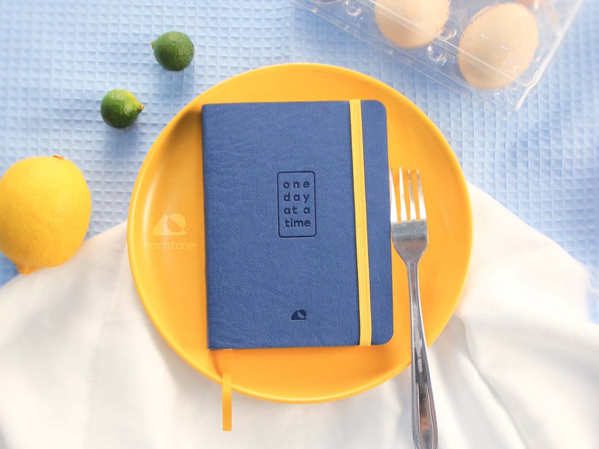 The O1 Journal, closed, on a yellow plate with a fork, beside a lemon, two limes, and a carton of eggs on blue cloth.