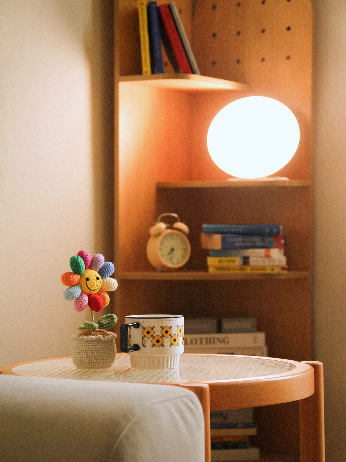The lamp lit on a wooden shelf at home, a crochet flower and a floral mug on the table in front.